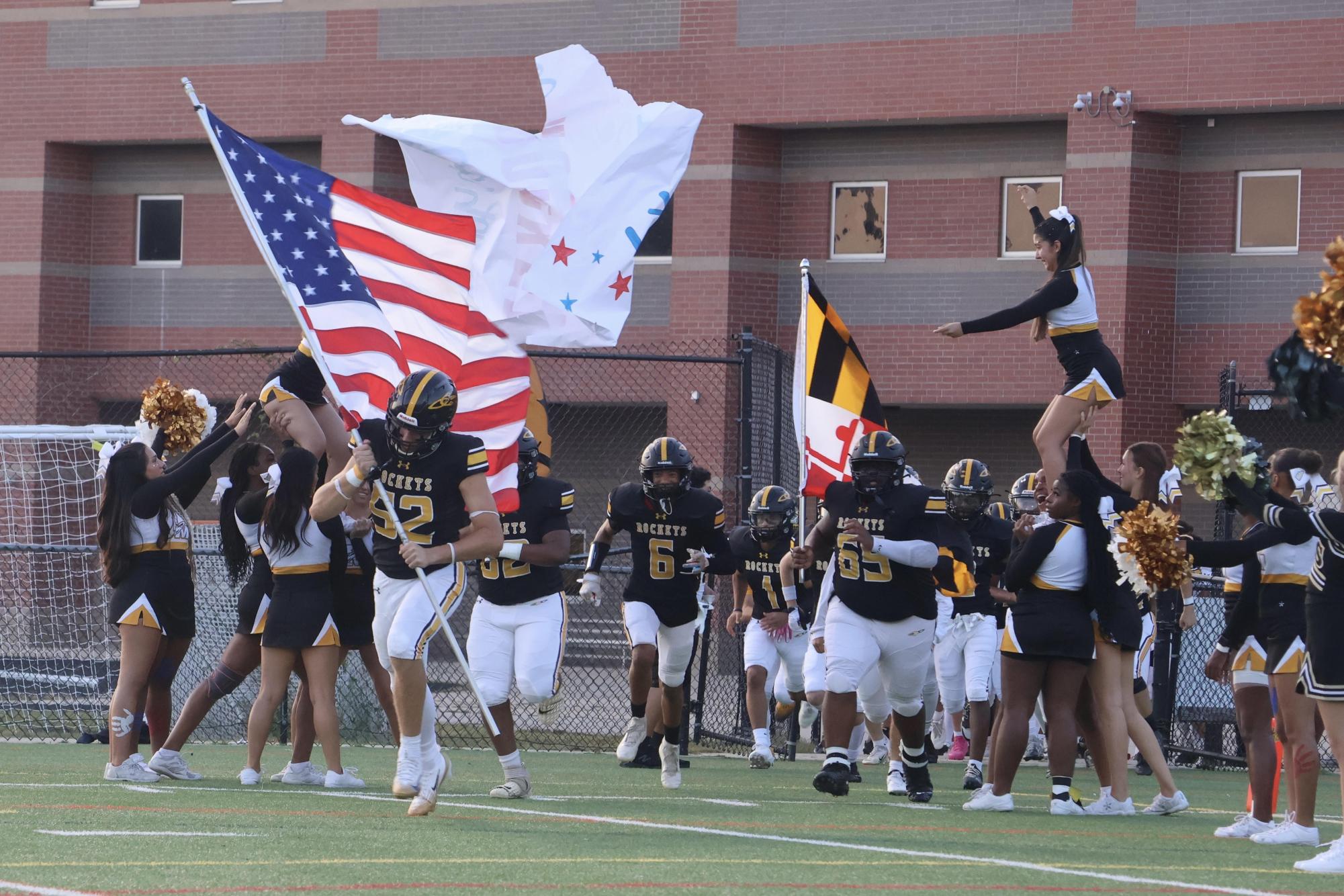 The football team runs through a banner carrying an American flag onto the field for their first home game of the season.