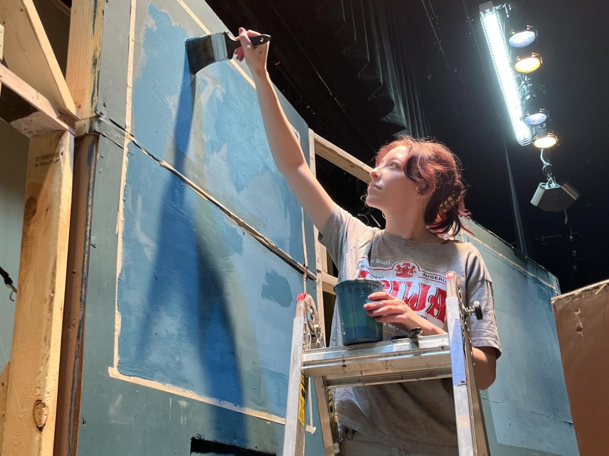 Brushing long navy stripes, senior and set decoration chief Kit Brown paints in the background for "The Play That Goes Wrong." 